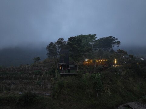 Kecamatan Getasan, Jawa Tengah, Indonesia - 08 September 2026: Aerial view of Merbabu View and Cafe on a misty hillside with warm string lights, lush trees, and terraced fields.