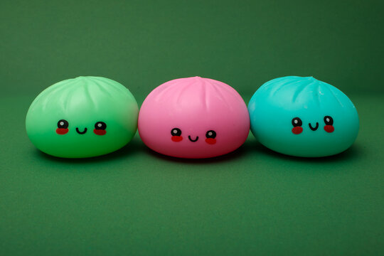 green, pink and teal kawaii squishy dumpling toys, one of each color, with cute smiley faces and rosy cheeks placed side by side on green table surface