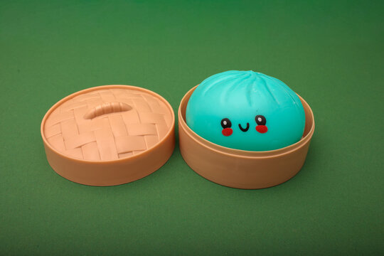 teal kawaii squishy dumpling toy in toy bamboo steamer basket with lid beside it on green background