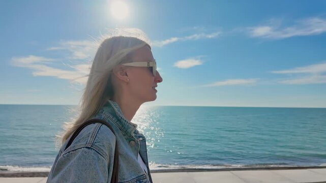 Side view: Attractive blonde woman in sunglasses and denim jacket walking along the promenade on a sunny day, the wind blowing her hair and revealing a beautiful seascape