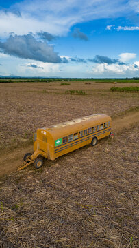 Boca de Yuma, La Altagracia Provinc, Dominican Republic - 12 September 2026: Aerial view of a modified yellow school bus trailer towed through a harvested field under a blue sky.