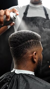 A professional barber applying hair spray to a young black man with a modern faded haircut in a salon shop.