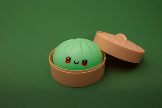 green kawaii squishy dumpling toy with cute smiley face and rosy cheeks placed on green background, soft silicone antistress toy for kids play, squeeze relief and adorable desk decoration