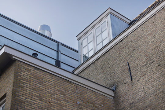 A white dormer window and galvanised roof trim stand against a clear blue sky, seen steeply from street level on an old brick facade.
