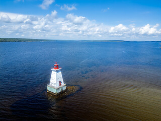 A red-and-white lighthouse stands on a rocky reef amidst vast blue coastal waters under a cloudy summer sky in Nova Scotia, Canada. © Margitta