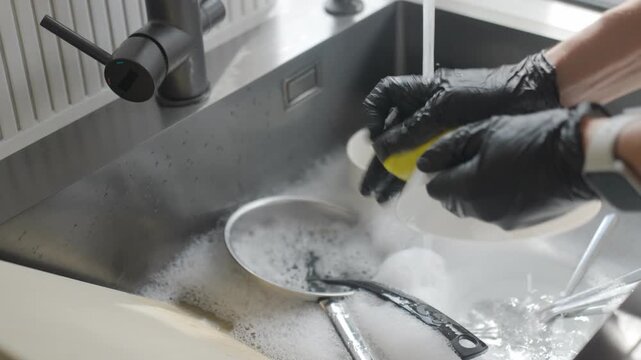 Woman in black cleaning gloves rinses and scrubs plate with sponge in soapy kitchen sink. Dishwashing, hand protection and everyday kitchen cleanup