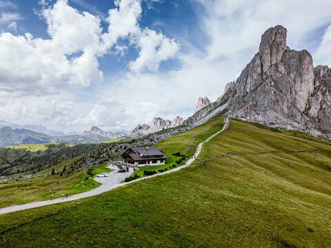 San Vito di Cadore, Italy - 21 August 2024: Aerial view of Giau Pass featuring a mountain hut nestled on grassy slopes with a winding path, rugged peaks, and a blue sky.