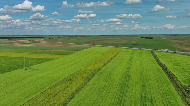 Aerial view of green agricultural fields and highway with trucks in Vojvodina plain, Serbia. Farmland landscape under blue sky clouds, 4k