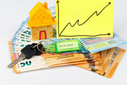 Yellow wooden toy house with a rising price chart note over fifty and twenty Euro banknotes with house keys on a white background