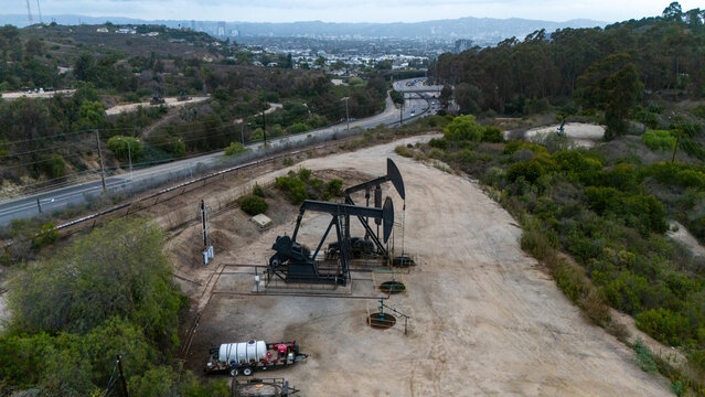 Aerial view of oil pump jacks in the Baldwin Hills area with surrounding hills, vegetation, and a highway in the distance in Los Angeles, California, United States.