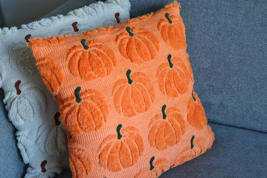 A bright orange pumpkin-patterned decorative pillow resting on a gray sofa. Concept of autumnal cozy home decor.