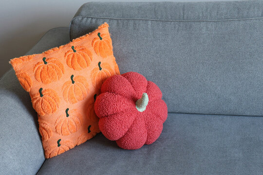 A coral pumpkin-shaped plush cushion rests against an embroidered pumpkin throw on a gray couch. Concept of seasonal texture and warmth.