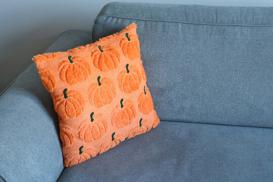 A bright orange pumpkin-motif cushion leans against the arm of a gray sofa. Concept of cheerful fall styling.