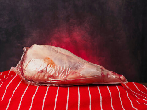 Raw lamb leg in plastic vacuum wrap rests on red and white table cloth, painted background. Meat shows pink flesh and red fat marbling. Scene suggests butchery, cooking, or grocery retail.