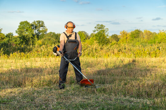 Man with brush cutter trims dry grass in sunny field. Protective earmuffs and gloves suggest focused yard work as maintenance worker stands ready in open meadow, feeling concentrated and calm.