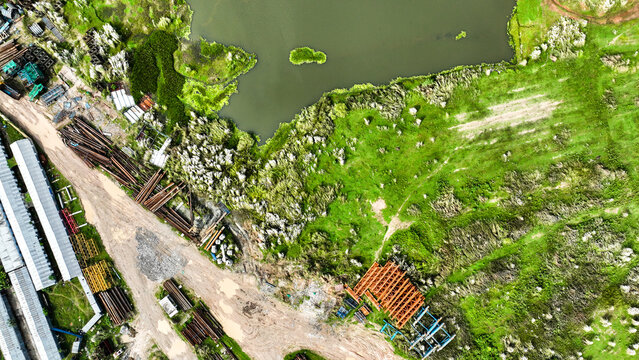 Aerial view of construction materials and metal pipes stored near a riverbank with lush green fields and dirt paths in Shariatpur, Dhaka Division, Bangladesh.