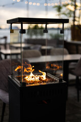 Fototapeta premium Modern outdoor gas fireplace with flames inside glass box for guests comfort at trendy restaurant patio, stylish gas heater with fire behind glass at cafe open-air terrace, cozy lounge settings.