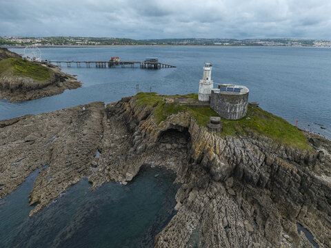 Aerial view of Mumbles Lighthouse situated on a rocky island with a pier and ferris wheel in the background under a cloudy sky in Swansea, Wales, United Kingdom.