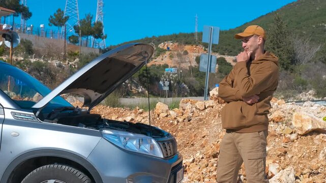 Frustrated man experiencing car trouble on a rural roadside. Confused man standing on the side of a country road, thoughtfully looking under the open hood of his broken-down car