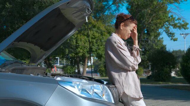 Frustrated woman examining broken car engine. Mature woman experiencing car trouble, holding her head in frustration while standing next to her vehicle with the hood open on a sunny day