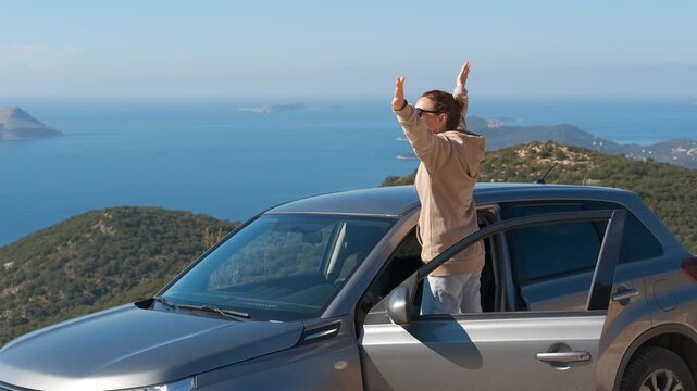 Free woman raising arms on a scenic road trip overlooking the ocean. Joyful woman getting out of her car and raising her arms in the air, celebrating freedom and success while enjoying the ocean view