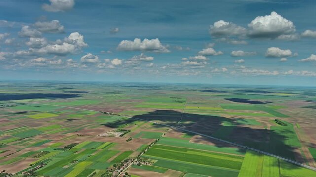 Aerial view of green farm fields stretching to horizon in Vojvodina plain. Patchwork farmland with village and trees under cloudy sky, 4k