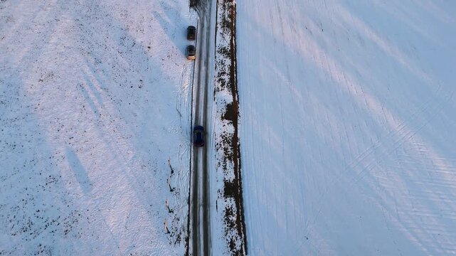 Aerial view to blue car driving along a snowy road bordered by trees and fields, showcasing the progression of travel through a winter landscape in a rural setting. Auto riding at rural route