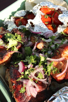 Grilled meat chunks garnished with red onion and cilantro, served with roasted tomatoes in foil under natural sunlight