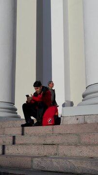 Man in Red Jacket Sitting in Front of a Building Looking at His Phone