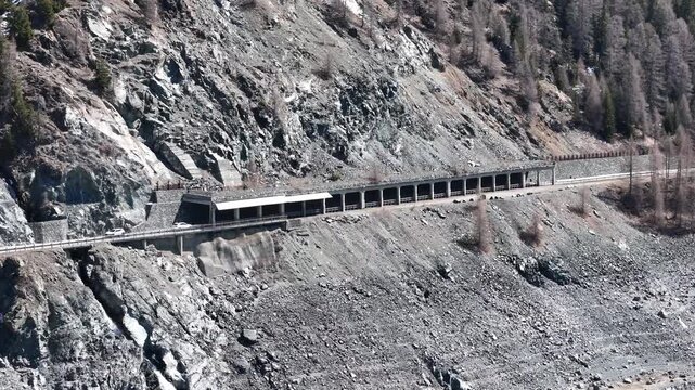 single car crosses julier pass road beside snow shed shielding steep scree slope waterfall cascades down rocky gully sparse larch trees dot mountainside aerial drone footage graubunden switzerland