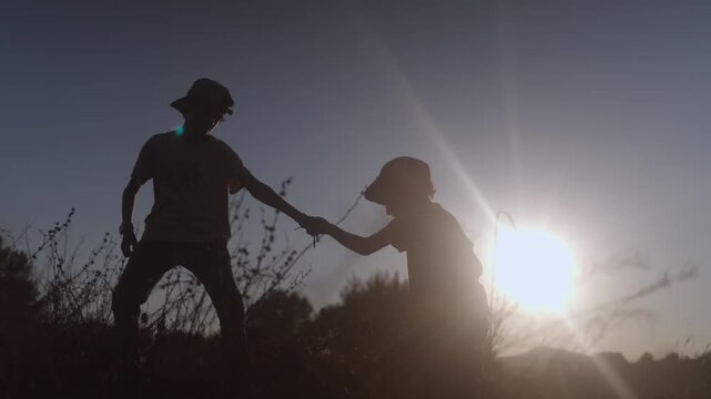 Silhouette of a boy helping his friend climb up a hill at sunset, symbolizing support, teamwork, friendship, and childhood memories in a beautiful natural setting