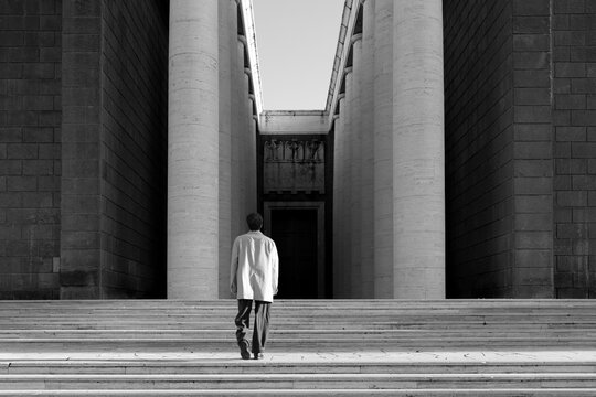 A man in a coat walks up wide stone stairs toward a monumental building with massive columns. Minimalist architectural spaces inspire deep reflections on human scale, ambition and authority.