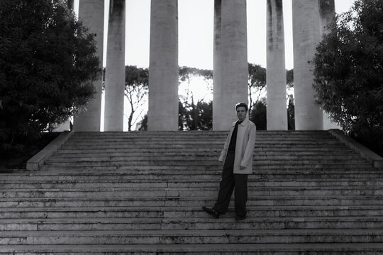 A young man in a coat looks back from monumental stone stairs in black and white. Classic architectural spaces inspire deep reflections on timeless elegance, personal journey and modern confidence.