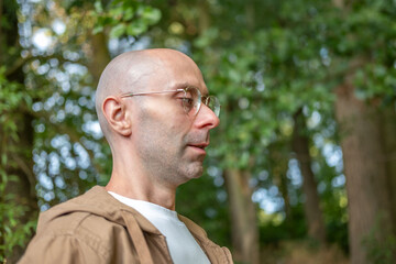 side profile portrait bald man glasses brown hoodie standing outdoors green park near trees, thoughtful Contemplative person enjoying peaceful nature, contemplative mood, nature scenery, peaceful day © kittyfly