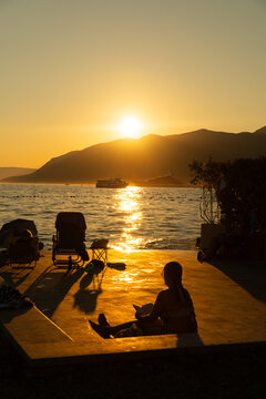 Tivat, Montenegro - 01 August 2025: View of a person reading on a terrace overlooking the sea at sunset with boats on the water and mountains in the background.