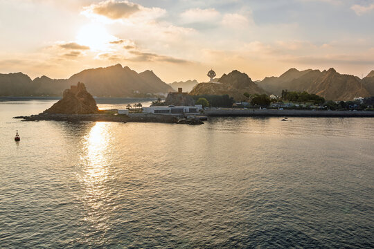 The Riyam Monument shaped like a giant incense burner rises above the scenic harbor and waterfront corniche with morning sun on the Al Hajar mountains behind, in Muscat Oman.