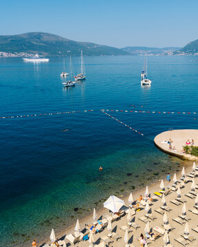 Tivat, Montenegro - 01 August 2025: View of the beach with rows of sun loungers and umbrellas, sailboats anchored in the clear blue water of the bay, and mountains in the background.