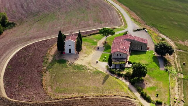 Aerial view of Chapel Vitaleta surrounded by rolling hills, cypress trees, and stone buildings in the rural landscape of San Quirico d'Orcia, Toscana, Italy.