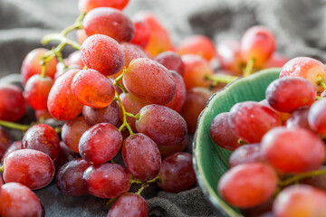 Close-up of fresh, glossy red grapes with water droplets, some on gray fabric and some in a green ceramic bowl. Vibrant ripe fruit ready to eat. © tai