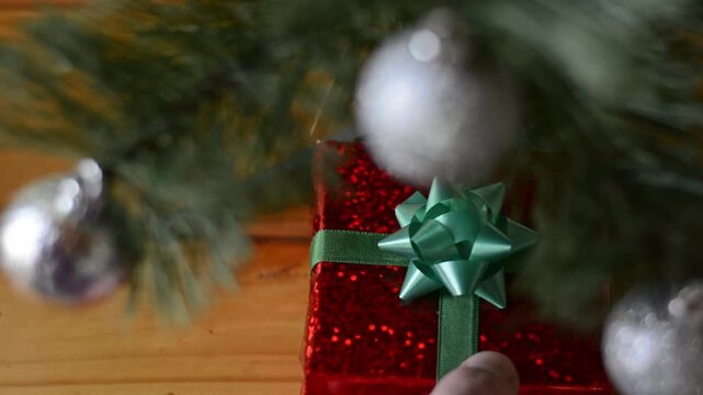 Top view of hand arranging wrapped gifts under Christmas tree with blurred tree branches and silver balls in the foreground, static camera
