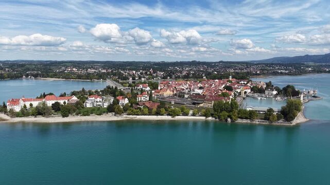 Aerial drone view of Lindau Island on Lake Constance in Bavaria, Germany, with the historic old town, harbor lighthouse, passenger boat and Alpine foothills under a dramatic cloudy sky.