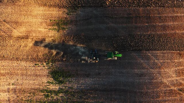 Aerial view of a green tractor plowing a field and creating dust in Val d'Orcia, San Quirico d'Orcia, Tuscany, Italy.