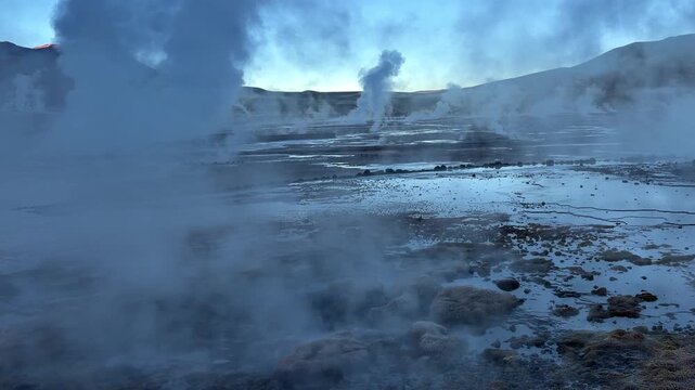 A geyser releases dense steam in the Atacama Desert. The site lies near Calama and Toconce in the Andes. Two shots show water vapour rising over arid stony ground.