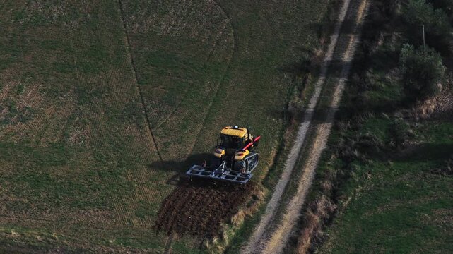 Aerial view of a yellow tractor plowing a green field with a dirt path nearby in San Quirico d'Orcia, Tuscany, Italy.