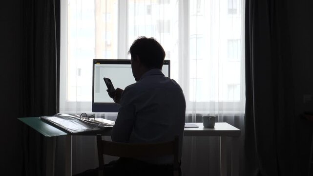 Man silhouette working at a desk by a window, using a computer and smartphone, representing concepts of remote work, productivity, technology, and modern professional life
