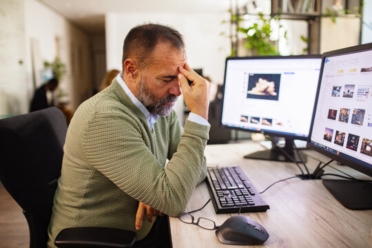 Stressed middle-aged man with headache at office desk using dual monitors