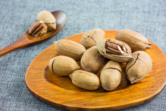 Shelled and in-shell pecans displayed on a wooden plate and spoon, set on a gray textured surface.