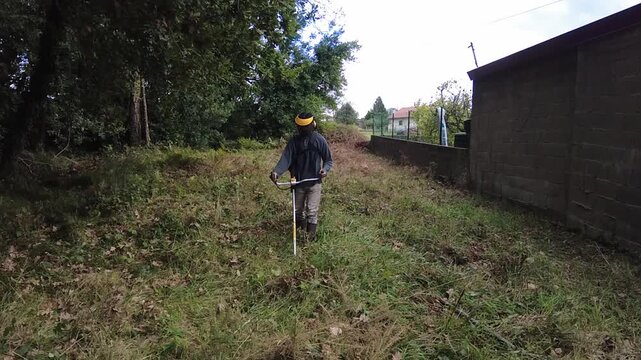Slow motion footage of a professional gardener using a powerful string trimmer to cut down overgrown grass and weeds in a backyard, performing routine seasonal landscaping maintenance work. 