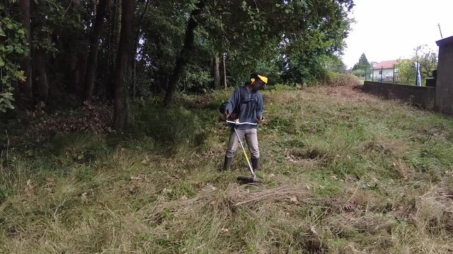 Slow motion footage of a professional gardener using a powerful string trimmer to cut down overgrown grass and weeds in a backyard, performing routine seasonal landscaping maintenance work. 