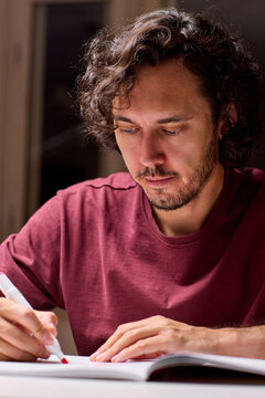 Focused adult man writing notes in open notebook at desk under warm lamp, seated portrait with maroon shirt, calm expression and concentrated posture